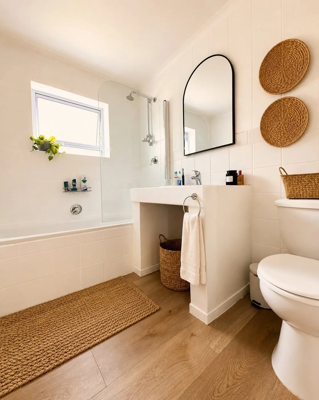 Van Hooch bathroom — white tiles, a black arched mirror, woven wall discs, a seagrass basket and a jute bath mat over oak-look flooring