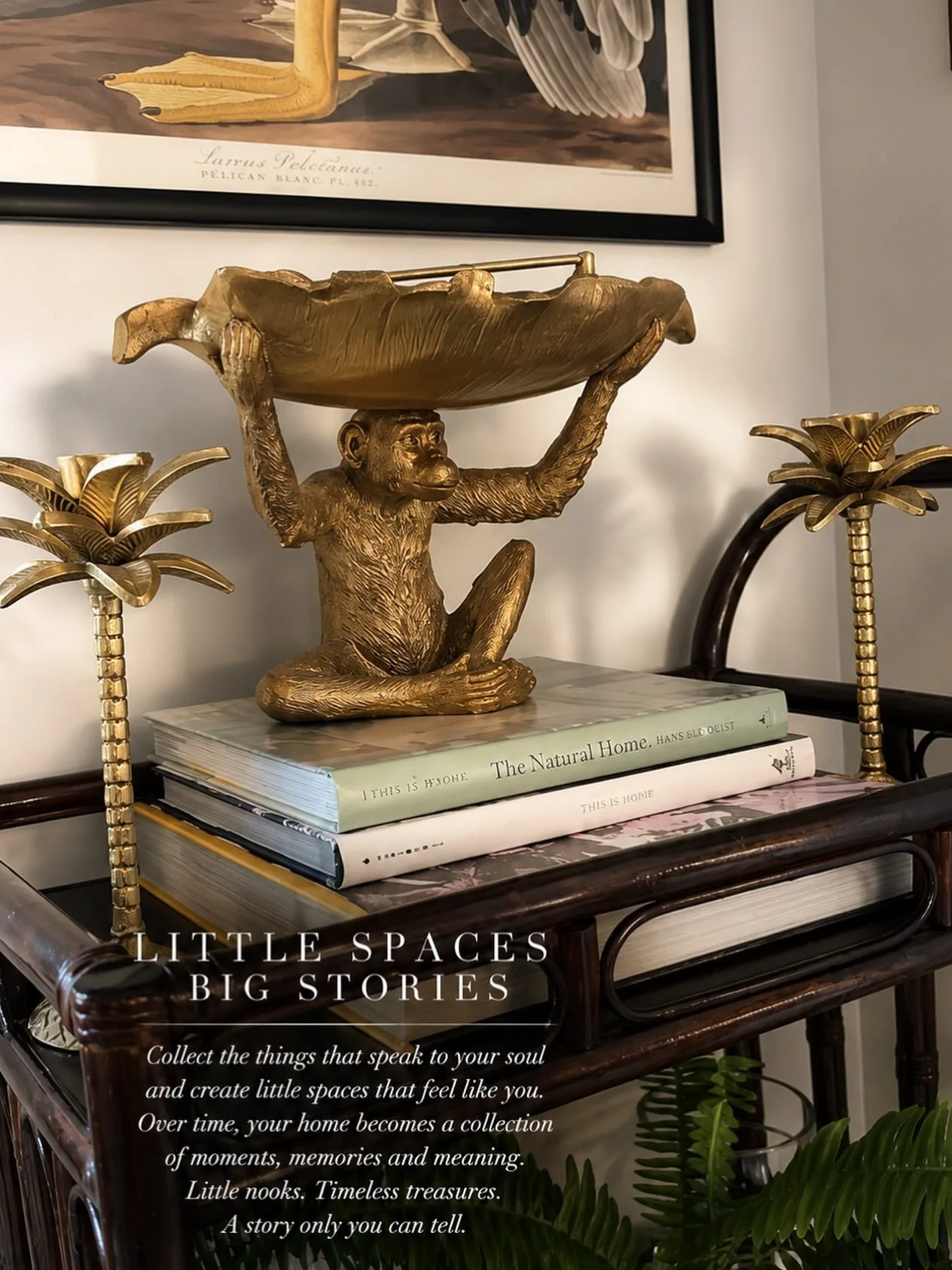 Van Hooch styling vignette — a brass monkey-and-leaf bowl on stacked coffee-table books, flanked by brass palm candlesticks beneath a framed pelican print